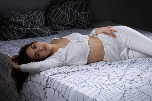 กสาวท าสนใจในช ดนอนส ขาวก อนบนเต ยงในห องนอนในแสงตอนเย — ภาพถ่ายสต็อก