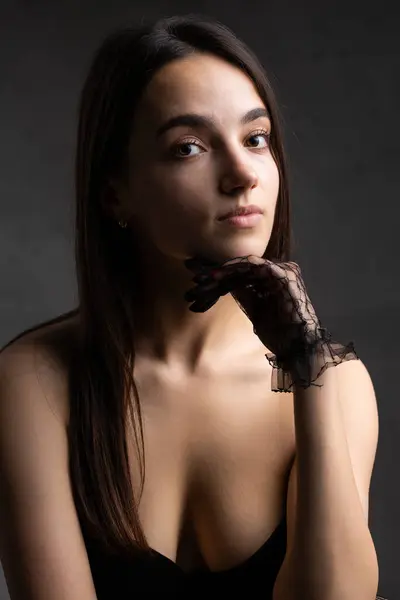 Classic dark studio portrait of a young brunette woman in black clothes who is sitting on a chair.