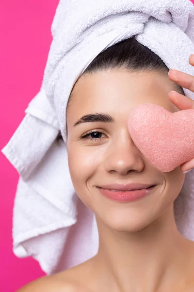 Beautiful cheerful attractive girl with a towel on her head, holds a sponge in the form of a pink heart.