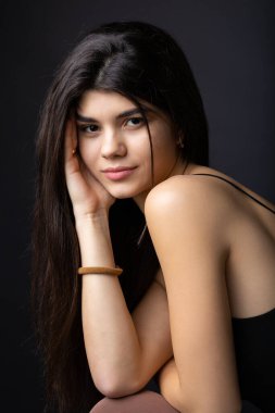 Classic studio portrait of a young brunette dressed in a black top, who is sitting on a chair against a black background.