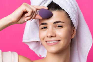 Beautiful cheerful attractive girl with a towel on her head, holds a sponge against a pink background.
