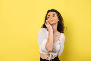 A young brunette girl with a cleansing scrub mask on her face, looks at the camera and smiles, against a yellow background.
