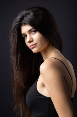Classic studio portrait of a young brunette dressed in a black top, who is sitting on a chair against a black background.