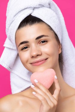 Beautiful cheerful attractive girl with a towel on her head, holds a sponge in the form of a pink heart.