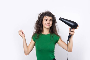 A cute young girl dressed in a green top dries her beautiful long silky hair with a hair dryer against a white background.