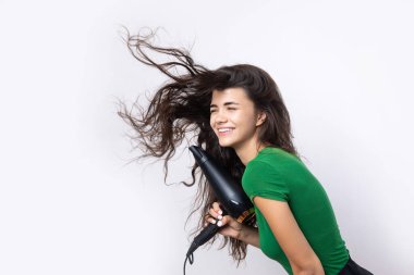 A cute young girl dressed in a green top dries her beautiful long silky hair with a hair dryer against a white background.