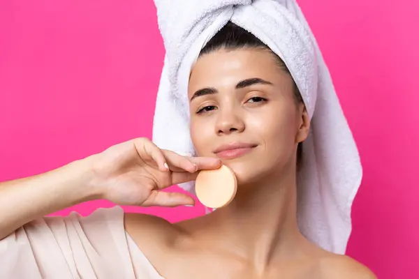 stock image Beautiful cheerful attractive girl with a towel on her head, holds a sponge against a pink background.