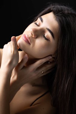 Classic studio portrait of a young brunette dressed in a black top, who is sitting on a chair against a black background.