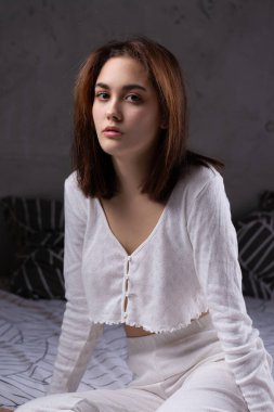 A young attractive girl dressed in white pajamas is resting on a bed in a bedroom in the evening lighting.