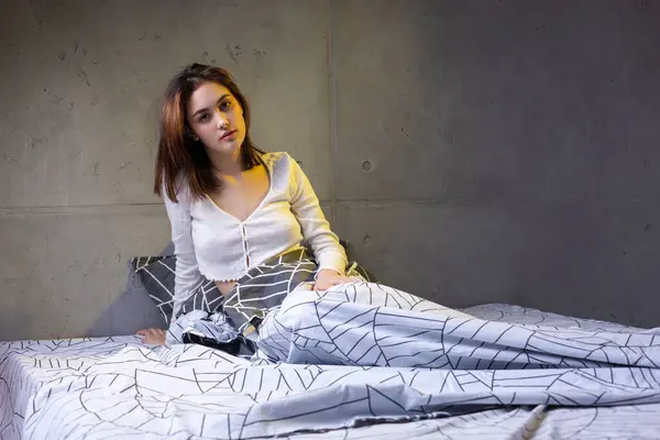 A young attractive girl dressed in white pajamas is resting on a bed in a bedroom in the evening lighting.