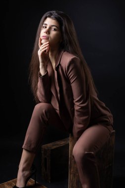 Classic studio portrait of a young brunette dressed in a black top and formal suit, who is sitting on a chair against a black background.