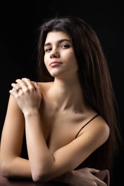 Classic studio portrait of a young brunette dressed in a black top, who is sitting on a chair against a black background.