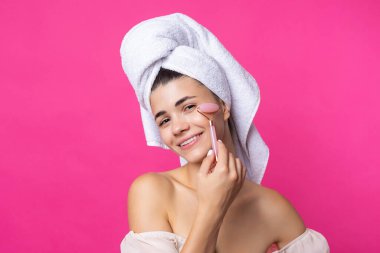 A beautiful cheerful attractive girl with a towel on her head holds a cosmetic roller near her face against a pink background.