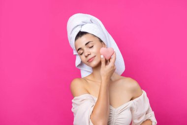 Beautiful cheerful attractive girl with a towel on her head, holds a sponge in the form of a pink heart.