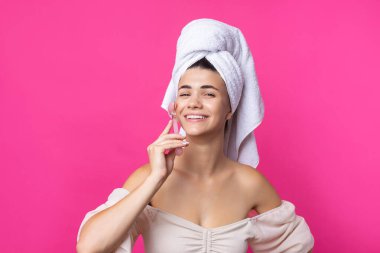 A beautiful cheerful attractive girl with a towel on her head holds a cosmetic roller near her face against a pink background.