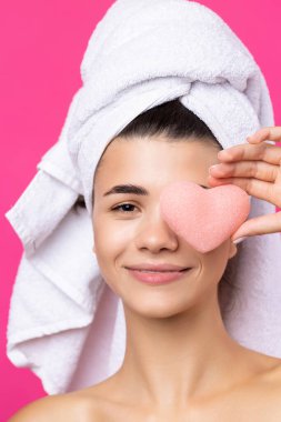 Beautiful cheerful attractive girl with a towel on her head, holds a sponge in the form of a pink heart.