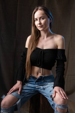 Classic studio portrait of a young brunette dressed in a black top, who is sitting on a chair against a black background.