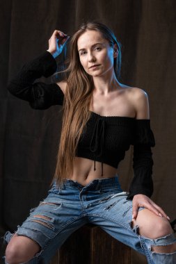 Classic studio portrait of a young brunette dressed in a black top, who is sitting on a chair against a black background.