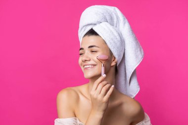 A beautiful cheerful attractive girl with a towel on her head holds a cosmetic roller near her face against a pink background.