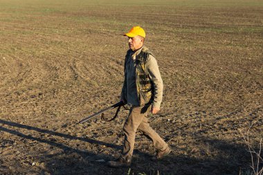 Mature man hunter with gun while walking on field.