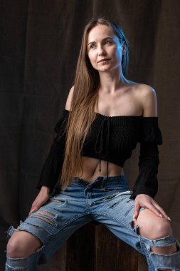 Classic studio portrait of a young brunette dressed in a black top, who is sitting on a chair against a black background.
