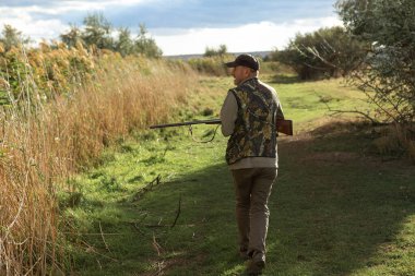 Mature man hunter with gun while walking on field.