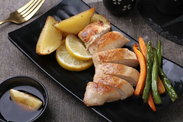 Kızarmış tavuk budu, patates ve siyah tabakta limon.