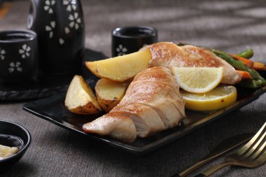 Ahşap masada siyah tabakta ızgara balık ve sebze.