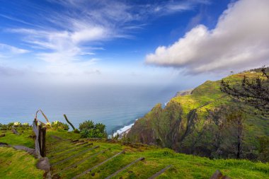 Madeira 'da (Miradouro dos Prazeres) güneşli bir günde yeşil bitkilerle kaplı bir tepenin dalgalarıyla kayalık sahili ve mavi Atlantik okyanusunu görmek
