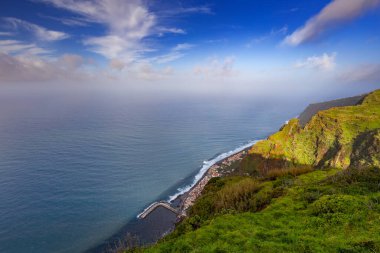 Madeira 'da (Miradouro dos Prazeres) güneşli bir günde yeşil bitkilerle kaplı bir tepenin dalgalarıyla kayalık sahili ve mavi Atlantik okyanusunu görmek