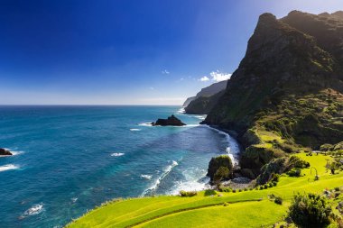 Madeira 'da (Miradouro de So Cristovo) güneşli bir günde yeşil çimlerle kaplı bir tepenin dalgalarıyla kayalık sahili ve mavi Atlantik okyanusunu görmek 