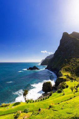 Madeira 'da (Miradouro de So Cristovo) güneşli bir günde yeşil çimlerle kaplı bir tepenin dalgalarıyla kayalık sahili ve mavi Atlantik okyanusunu görmek 
