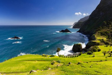 Madeira 'da (Miradouro de So Cristovo) güneşli bir günde yeşil çimlerle kaplı bir tepenin dalgalarıyla kayalık sahili ve mavi Atlantik okyanusunu görmek 