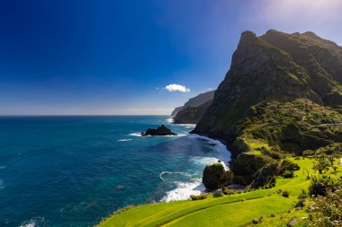 Madeira 'da (Miradouro de So Cristovo) güneşli bir günde yeşil çimlerle kaplı bir tepenin dalgalarıyla kayalık sahili ve mavi Atlantik okyanusunu görmek 