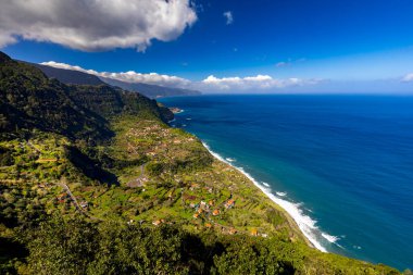 Madeira 'da güneşli bir günde (Miradouro da Beira da Quinta) yeşil bitkilerle kaplı bir tepenin dalgalarıyla kayalık sahili ve mavi Atlantik okyanusunu görmek, arka plan veya duvar kağıdı