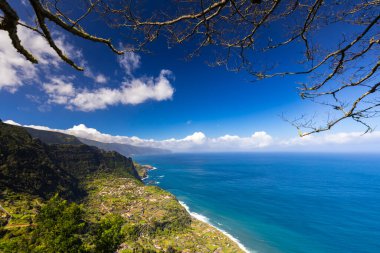 Madeira 'da güneşli bir günde (Miradouro da Beira da Quinta) yeşil bitkilerle kaplı bir tepenin dalgalarıyla kayalık sahili ve mavi Atlantik okyanusunu görmek, arka plan veya duvar kağıdı