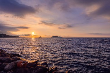 A picturesque seaside landscape at sunset with calm water, lush vegetation and mountains under a clear sky
