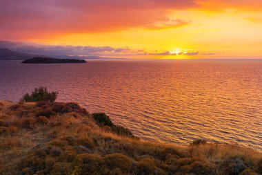 A picturesque seaside landscape at sunset with calm water, lush vegetation and mountains under a clear sky