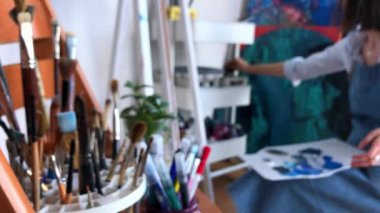 Stüdyosunda çalışan bir kadın sanatçı. Tüplerden beyaz palete mavi, beyaz ve siyah akrilik boya sıkıyor. Enstrüman standının üzerinde bulanık bir görüntü. Yüksek kalite 4K görüntü.