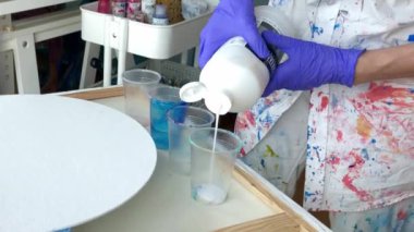 Kadın sanatçı akrilik boya karıştırıyor. Büyük şişeden plastik bardaklara kadar beyaz akrilik boya döken mor lastik eldiven ve renkli gömlek giyen bir kadın sanatçının ellerine yaklaş. Yüksek kalite 2K görüntü.