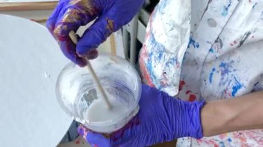 Kadın sanatçı akrilik boya karıştırıyor. Plastik bir bardağa beyaz akrilik boya karıştıran mor lastik eldiven ve renkli gömlek giyen bir kadın sanatçının ellerine yaklaş. Yüksek kalite 2K görüntü.