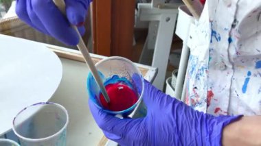 Kadın sanatçı akrilik boya karıştırıyor. Plastik bir fincanda kırmızı akrilik boya karıştıran mor lastik eldiven ve renkli gömlek giyen bir kadın sanatçının ellerine yaklaş. Yüksek kalite 2K görüntü.