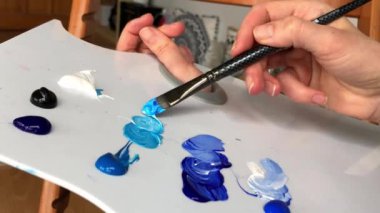 Beyaz palette akrilik boya karışımı. Mavi, beyaz ve siyah akrilik boyayı beyaz palette fırçayla karıştıran bir kadın sanatçının ellerine yaklaş. Yüksek kalite 2K görüntü.