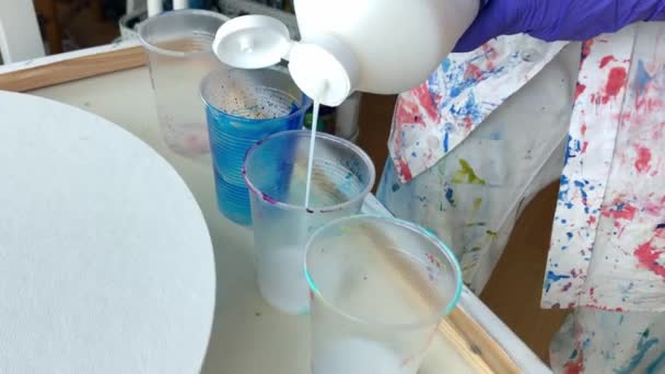 アクリル塗料を混ぜた女性アーティスト ヴァイオレットゴムグローブを着用した女性アーティストの手に近づき 大きなボトルからプラスチックカップに白いアクリルペイントを注ぐカラフルなステンドシャツ — ストック動画