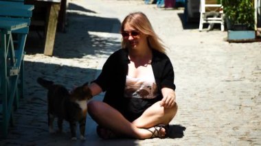 Siyah mayolu, güneş gözlüklü ve şapkalı genç sarışın kız sokağın kıyısındaki iskelede oturuyor.