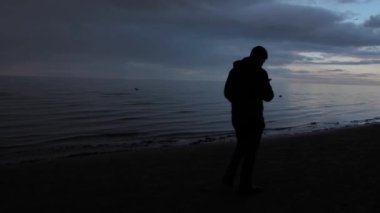 Alacakaranlıkta deniz kenarında yalnız bir adam duruyor, telefonuna dalmış akşam güneşinin altında..