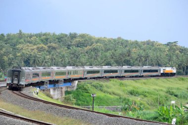 Kırsal ya da tepelik bir alanda hareket eden bir yolcu treni görüntüsü. İki tren hattı ve güzel manzarası var..