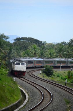 Kırsal ya da tepelik bir alanda hareket eden bir yolcu treni görüntüsü. İki tren hattı ve güzel manzarası var..