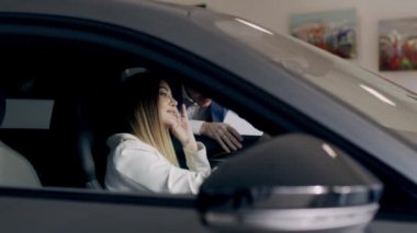 Genç ve güzel bir kız araba galerisinde yeni bir elektrikli araba seçiyor. Kadın mutlu, gülümseyen ve yeni bir araba almaktan memnun. Kız yeni arabadan memnun kaldı..