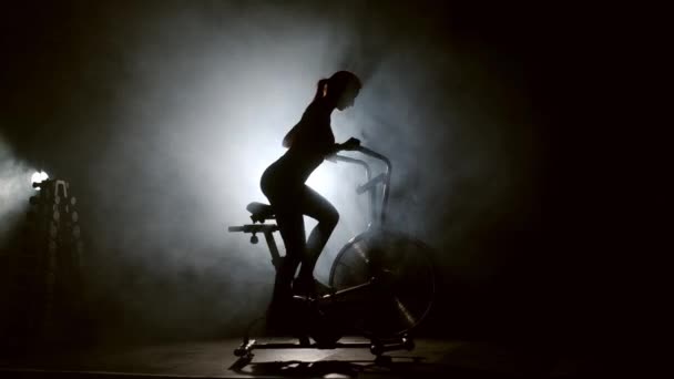 在现代的黑暗体育馆里 女运动员正在使用运动器材 一个年轻的女人在烟雾中骑自行车 在一个黑暗的健身房里对一个固定的人进行积极的肌肉教育 健康的生活方式 — 图库视频影像
